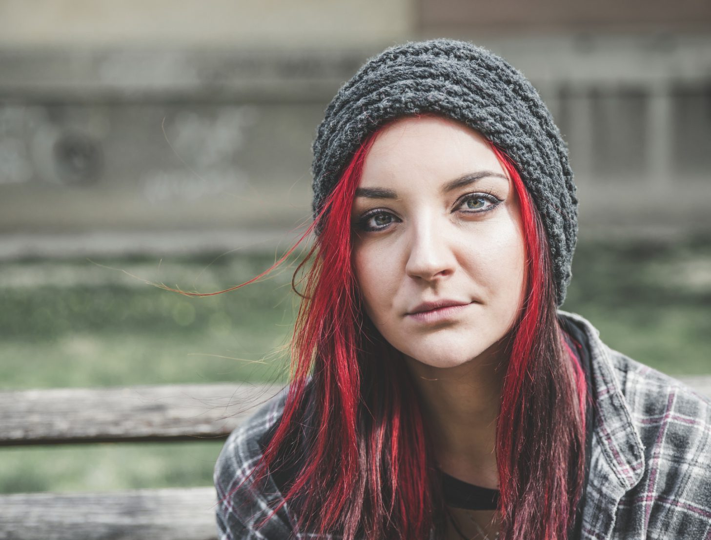 the photo shows a young girl sitting outside on a park bench, she has green eyes and long straight red hair, she is looking at the camera with a neutral expression