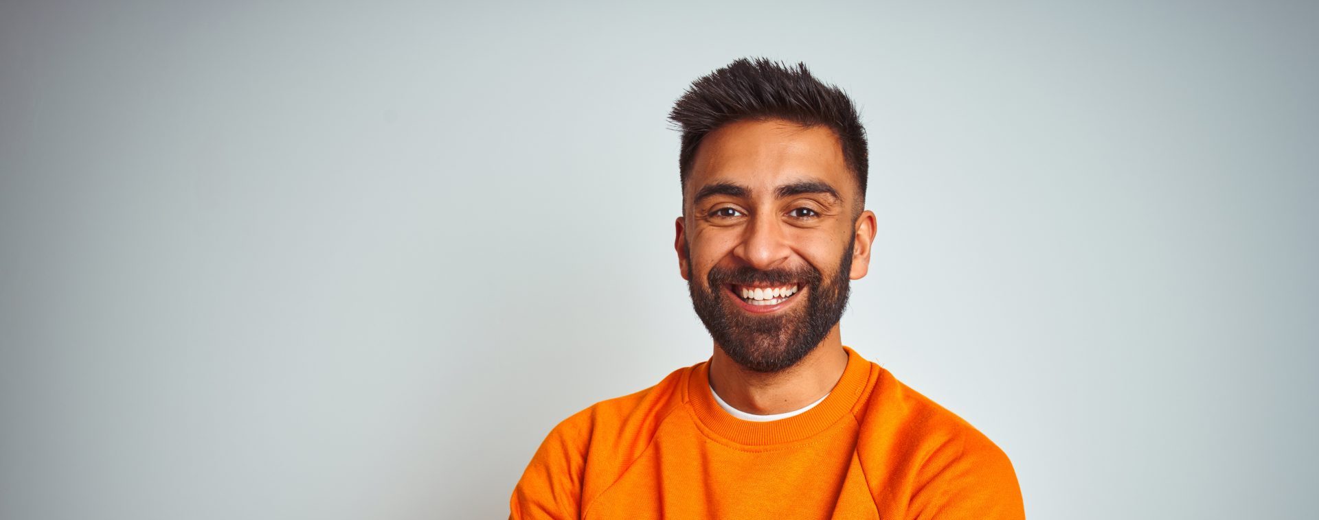a young man with dark hair, eyes and a beard is standing inside and smiling at the camera with his arms folded